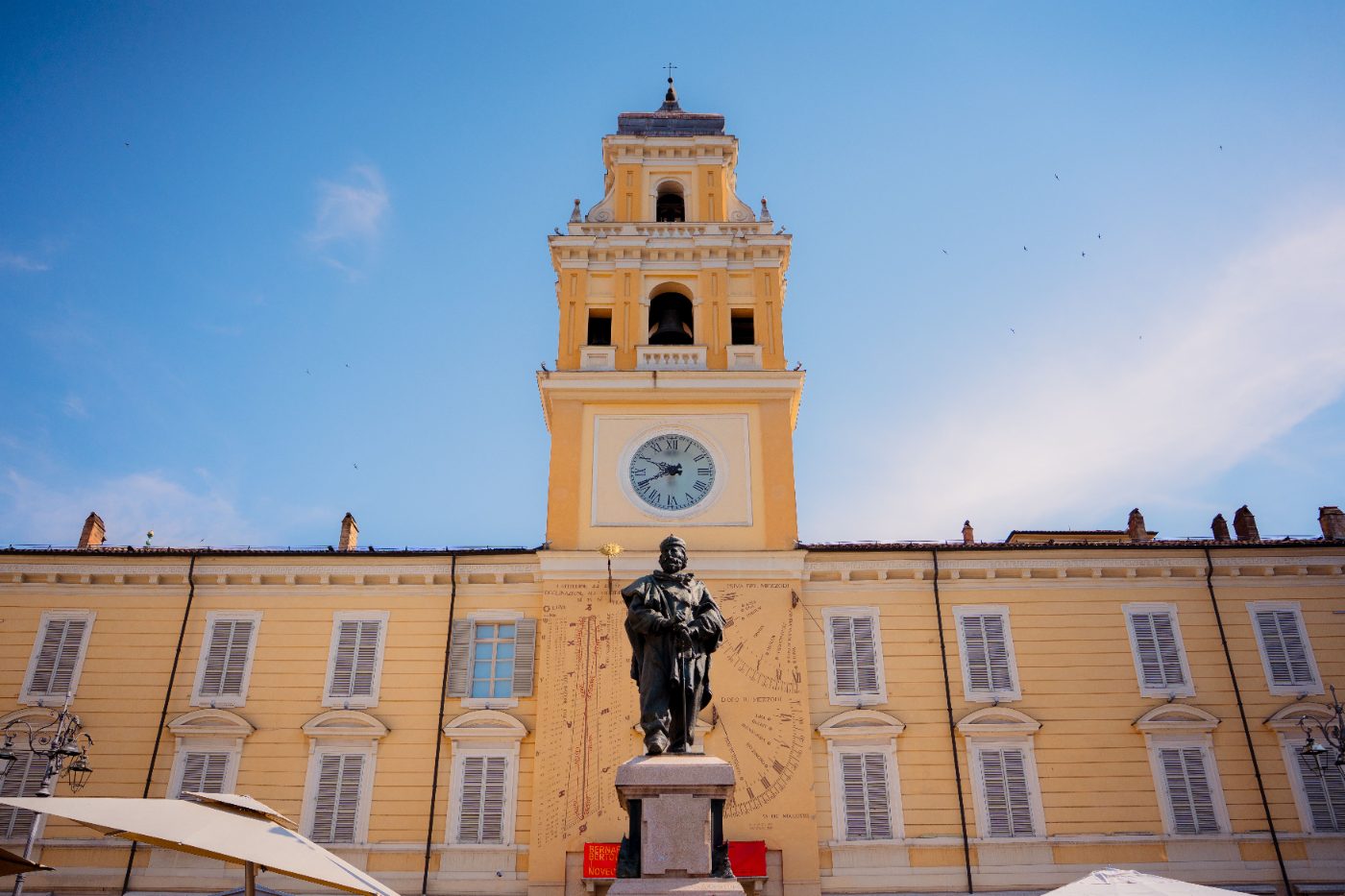 A historic clock tower and statue under a clear blue sky in Parma, Italy.